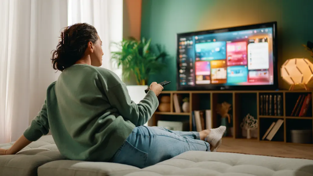 Lifestyle image of a woman in a green sweater sitting on a couch and holding a remote pointed at a TV