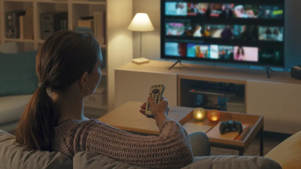 Lifestyle image of woman on a couch holding a remote pointed at a TV