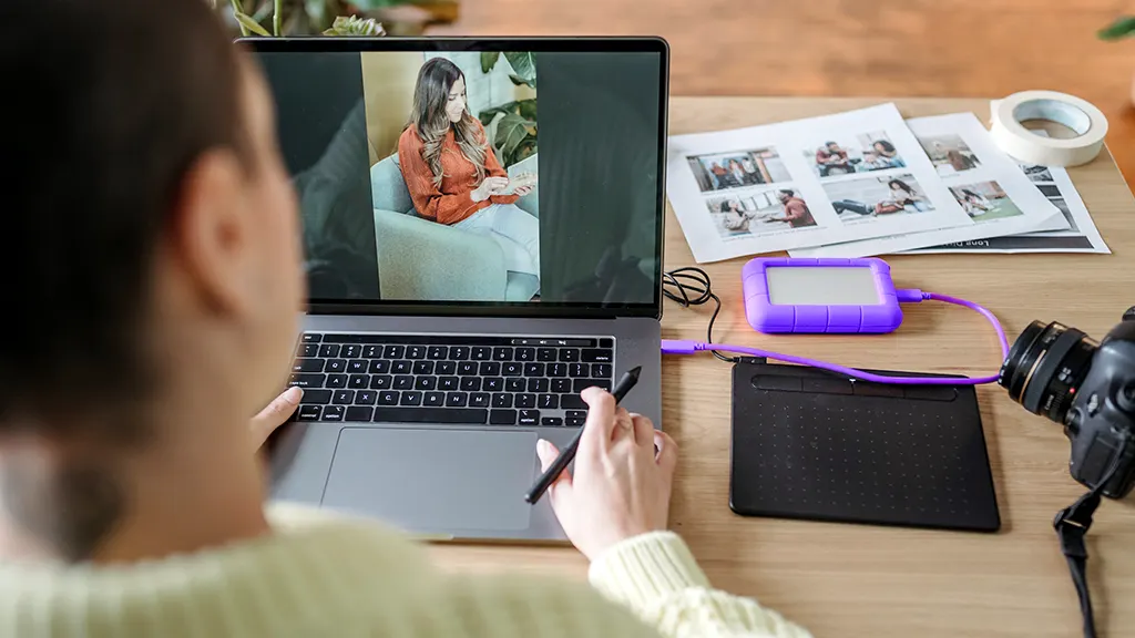 A woman sits pensively, facing her laptop screen, debating silently whether to move the image of another woman, looking at a phone screen, to an attached portable hard drive.