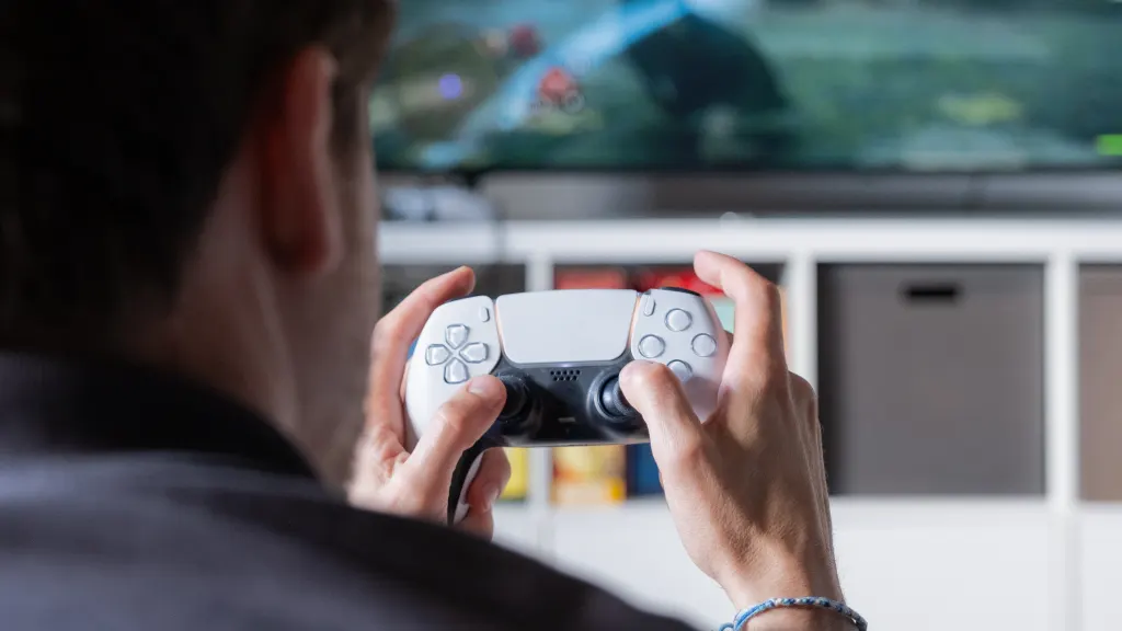 Close-up of a man holding a Playstation 5 controller and looking at a TV screen