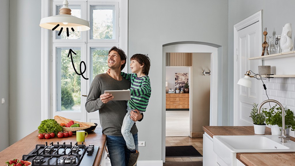 Father holding son in kitchen, controlling smart lightbulb from a tablet