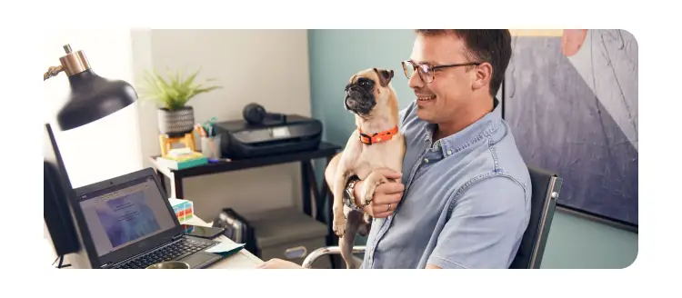 Professional working from a home office while holding a small dog, showcasing a productive remote work setup with a laptop and comfortable workspace.