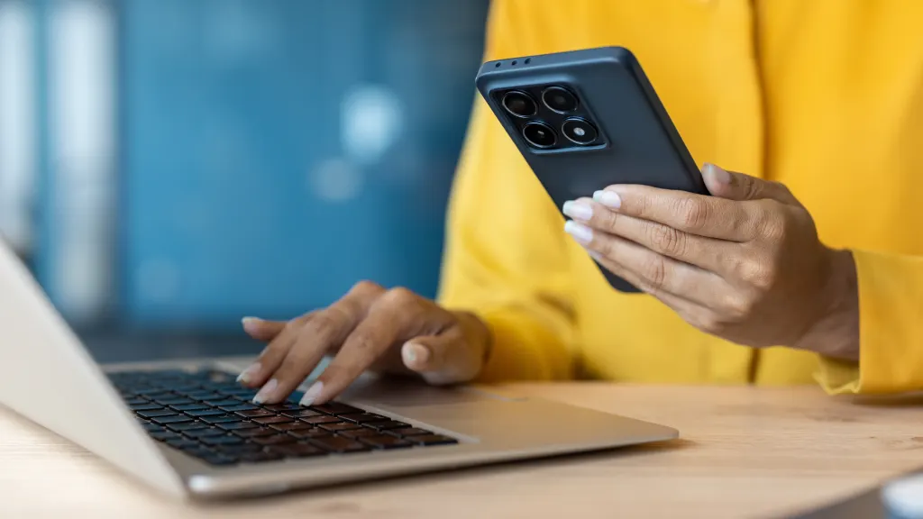 Closeup of person in a yellow sweater holding a phone and typing on a laptop
