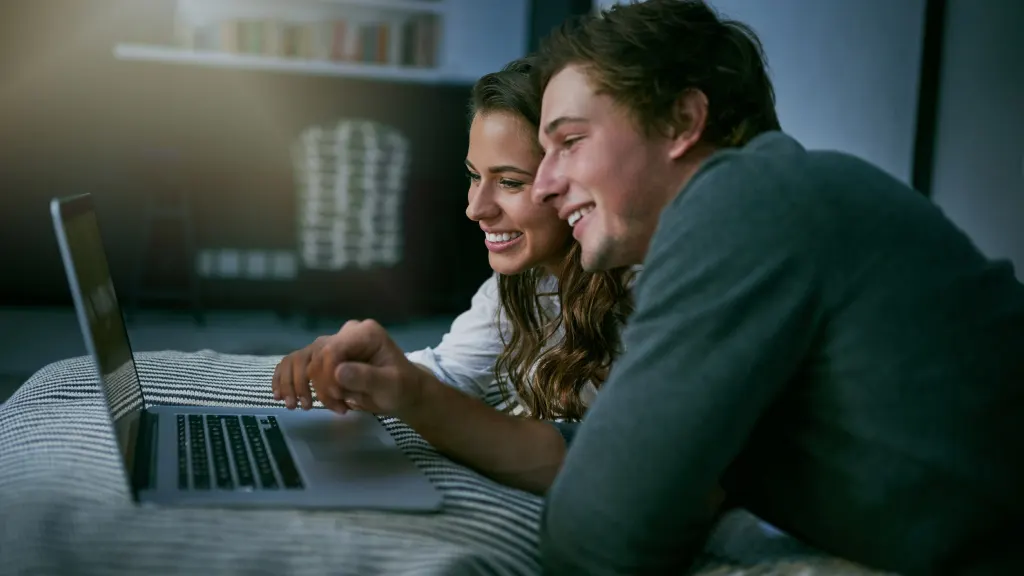 Lifestyle image of a couple laying on a bed and looking at a laptop