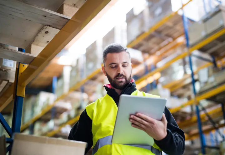 Warehouse employee in safety vest checking inventory on a tablet in a storage facility.