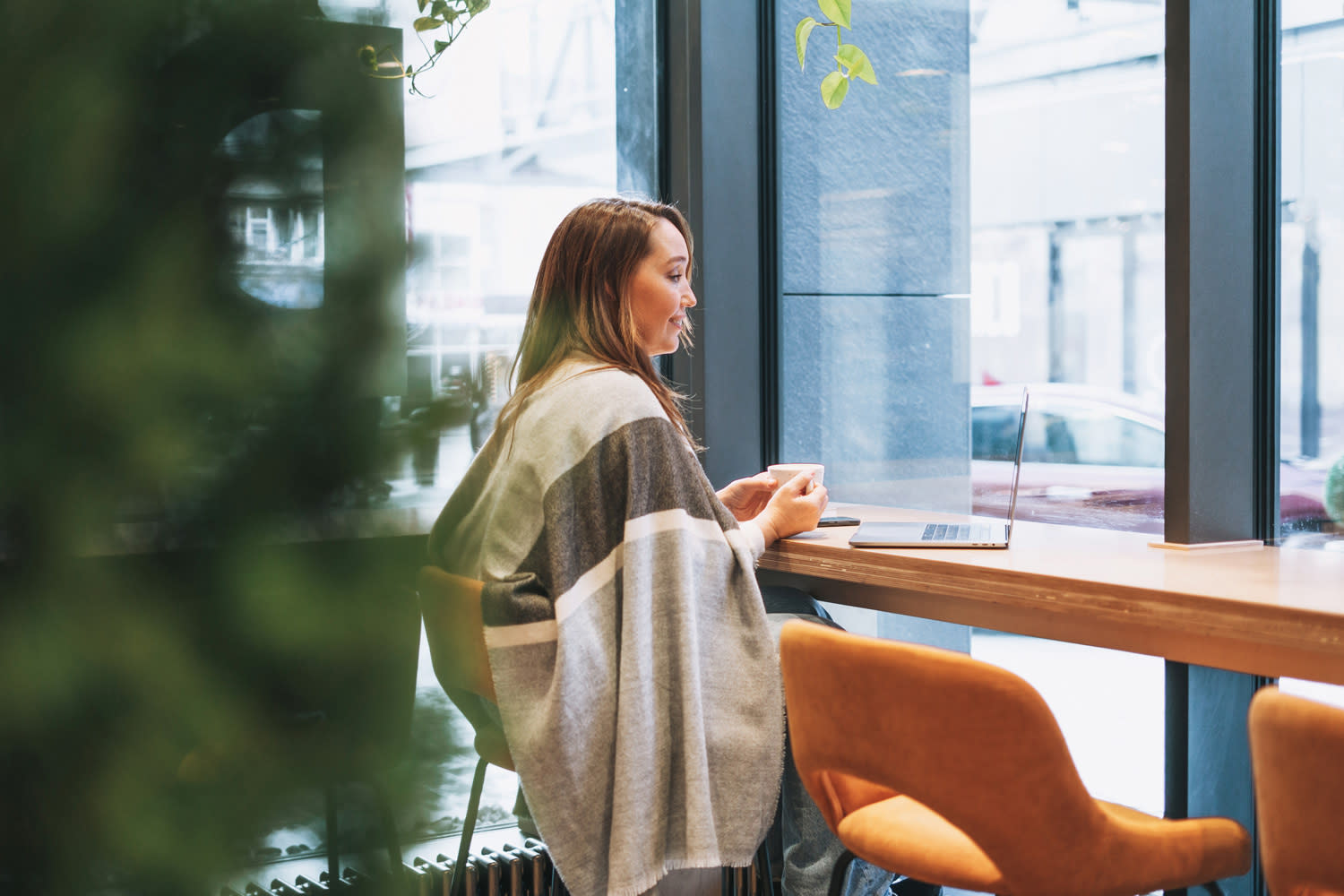 Kvinne sitter på cafe med kaffe og laptop. Ser tenkende ut.