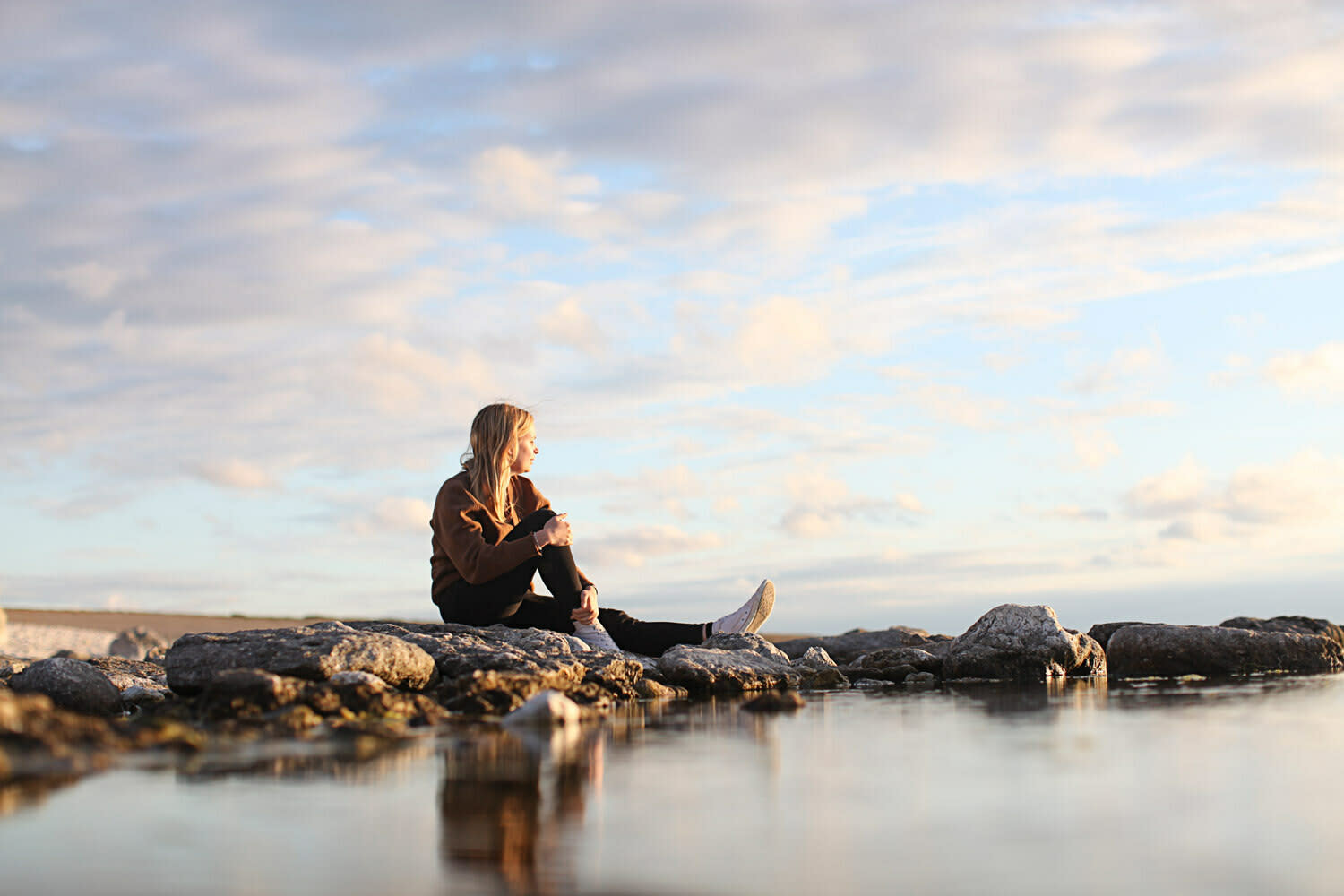 Blond kvinne sitter i strandkanten og ser utover havet