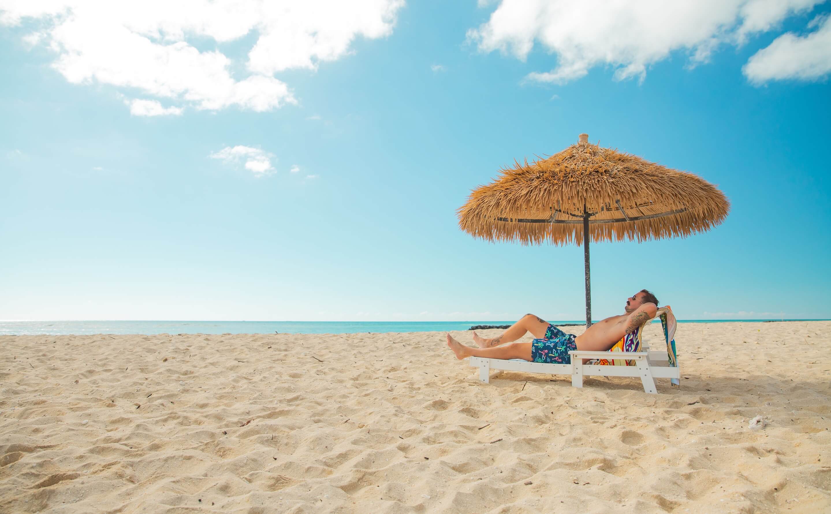 A man lounges in a beach chair under an umbrella. He’s wearing bright, floral printed Chubbies.