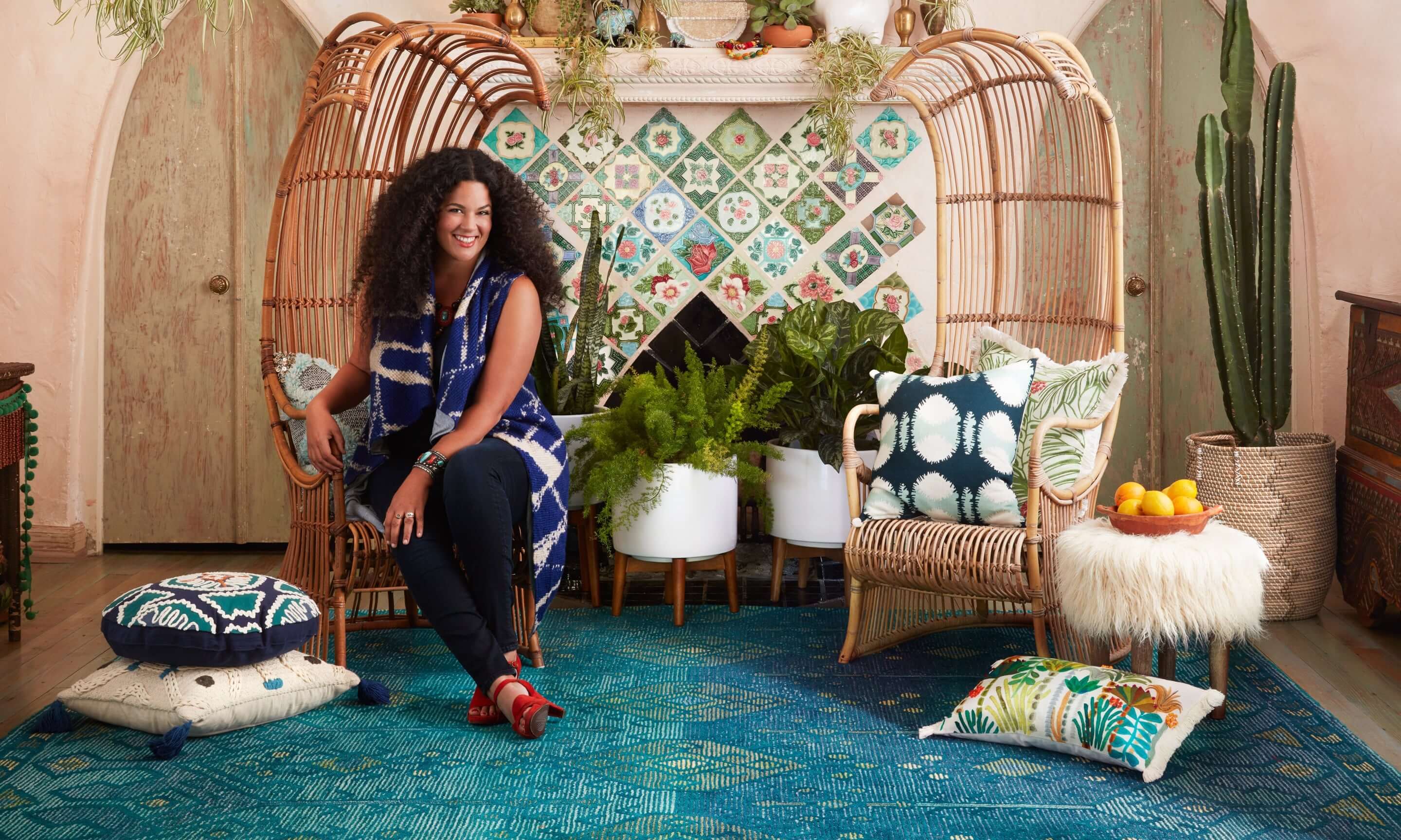 Founder of The Jungalow, Justina Blakeney, sits in a tall wooden chair in a room nicely decorated with many colourful pillows and an assortment of decorative potted plants.