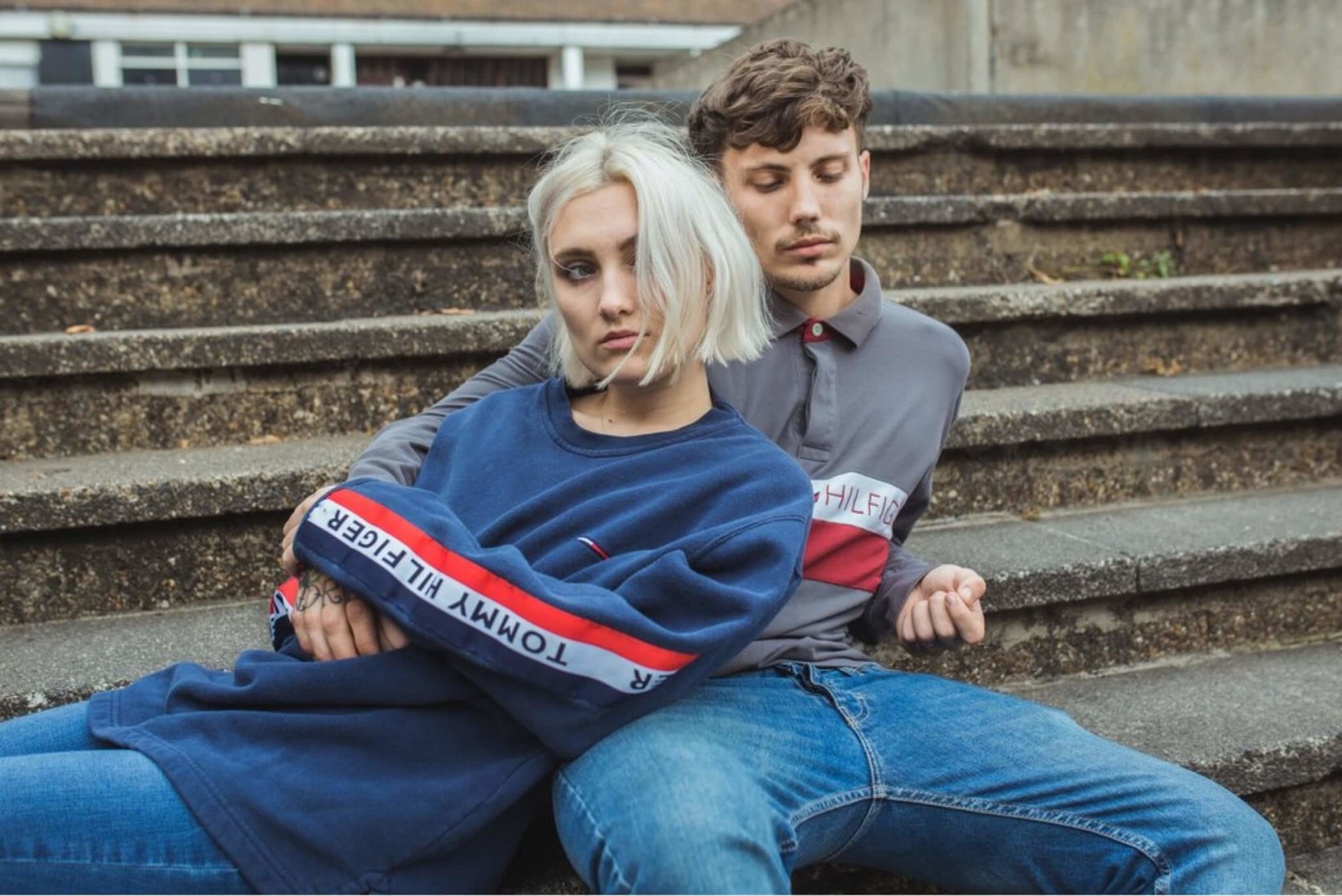 A woman lounges, arms crossed, on a male companion, seated on concrete stairs modelling a vintage Tommy Hilfiger sweater.