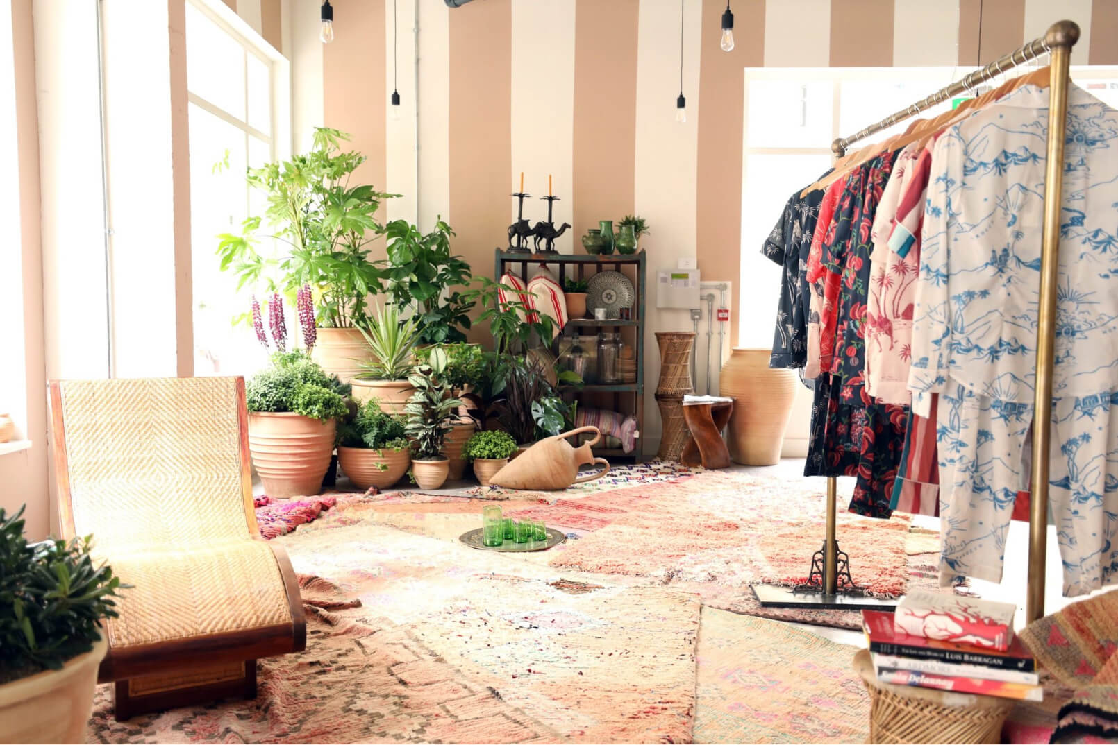 A golden clothing rack with several pairs of colourful Desmond & Dempsey pyjamas, hanging in a bright room with many plants.