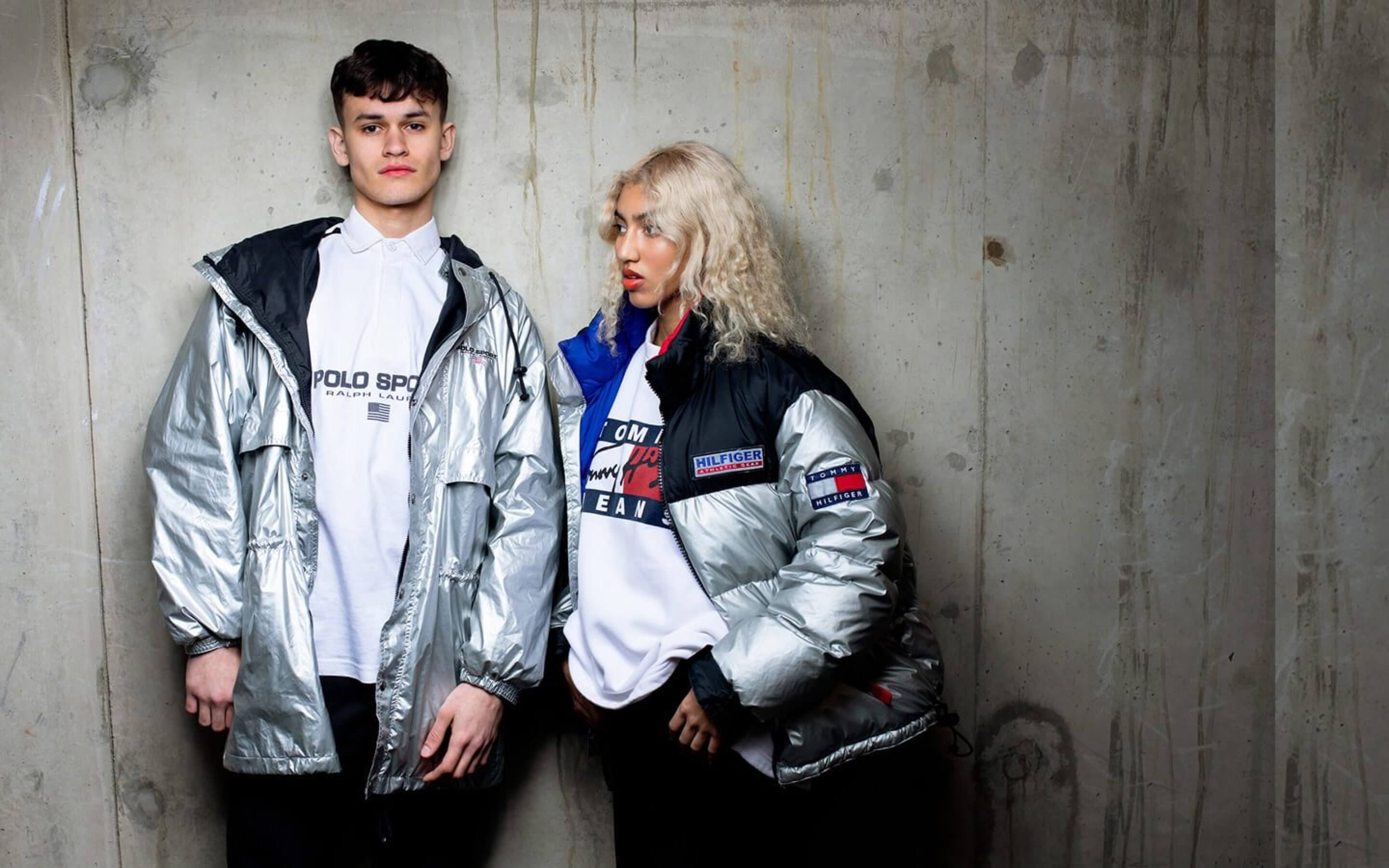 A male and female model stand against a concrete backdrop, dressed in white vintage sweaters and metallic grey vintage jackets.