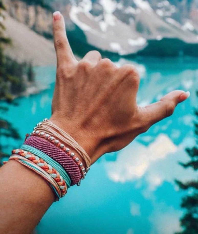 A woman’s hand wearing several Pura Vida bracelets making the shaka brah hand sign infront of a bright blue lake.