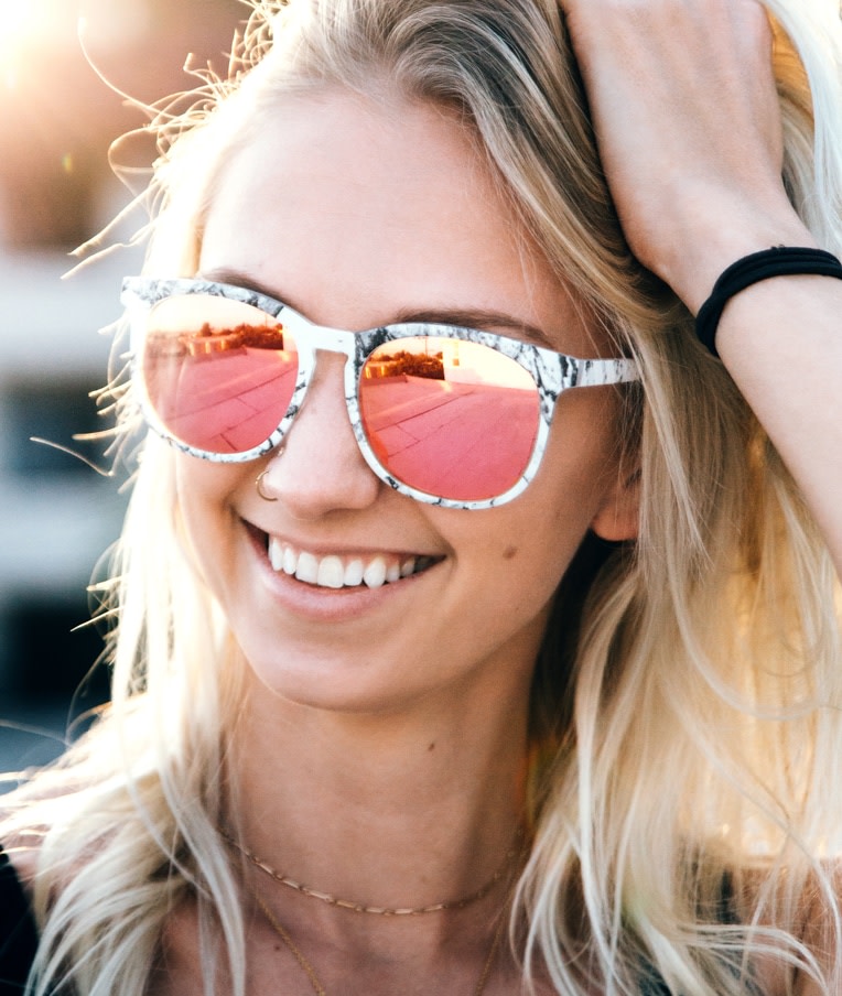 A smiling woman, she’s wearing a pair of white sunglasses with black marbling and a pinkish yellow tint.