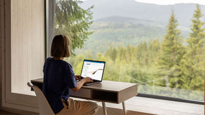 A woman sits at a desk in front of a laptop, with a window view of a forest in front of her