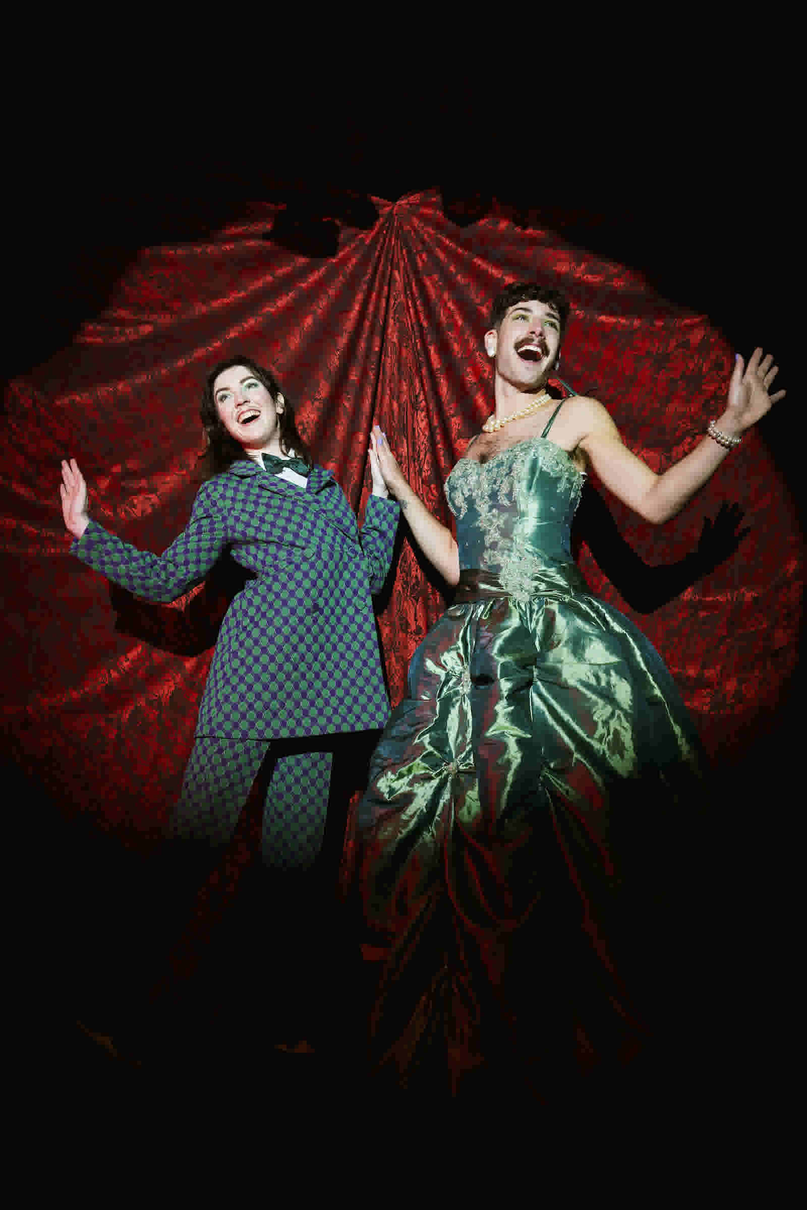 Two non-binary people pose in a spotlight showing over exaggerated delight. One is femme-presenting wearing a suit. The other is male-presenting wearing a dress. 