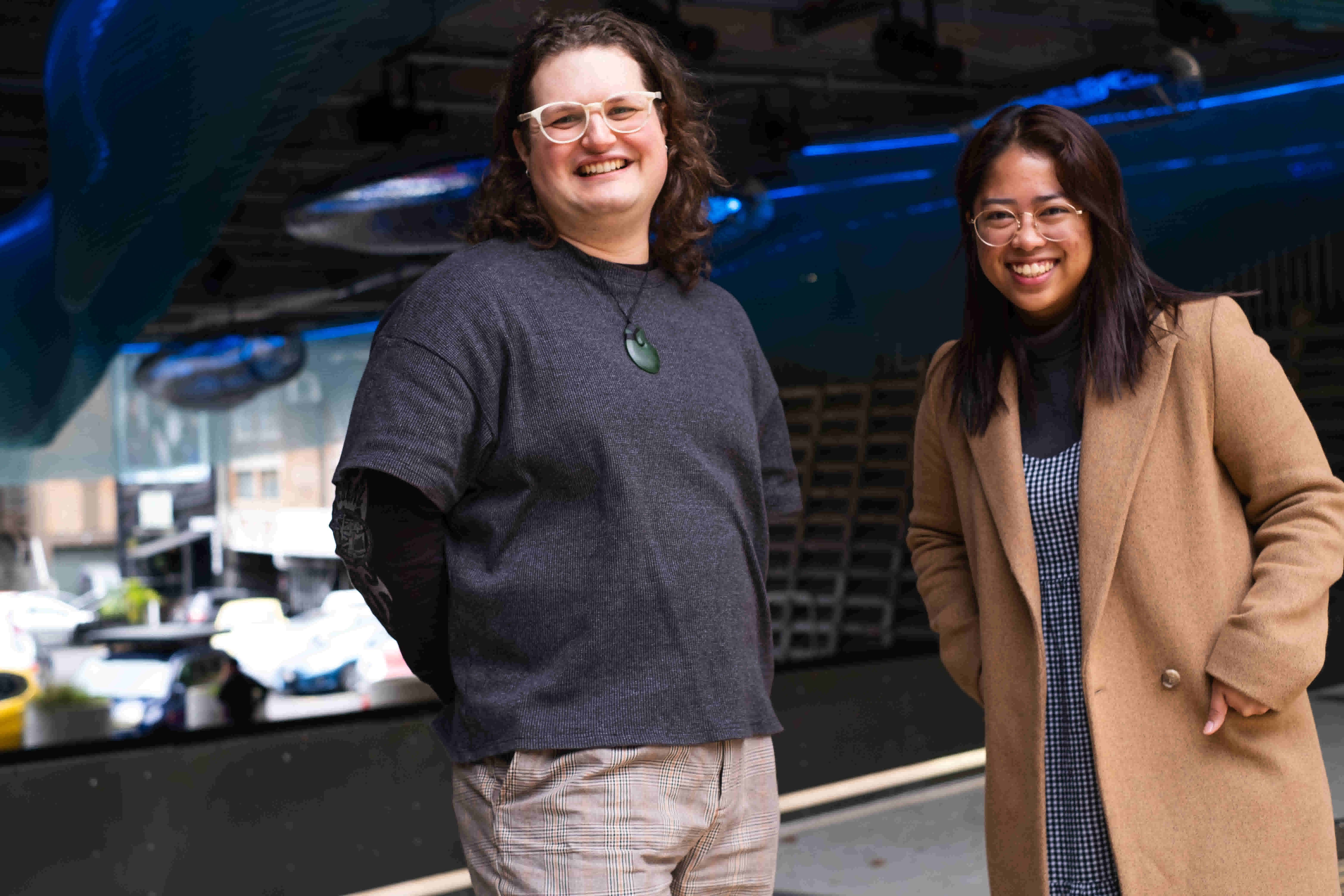 Image Description: Izzy and Alyssa stand next to each other with joyful expressions. They are standing underneath Waimahara, an interactive artwork with aqua-blue lights of lines and spirals encapsulated within a netting formed into tubular and wave shapes.