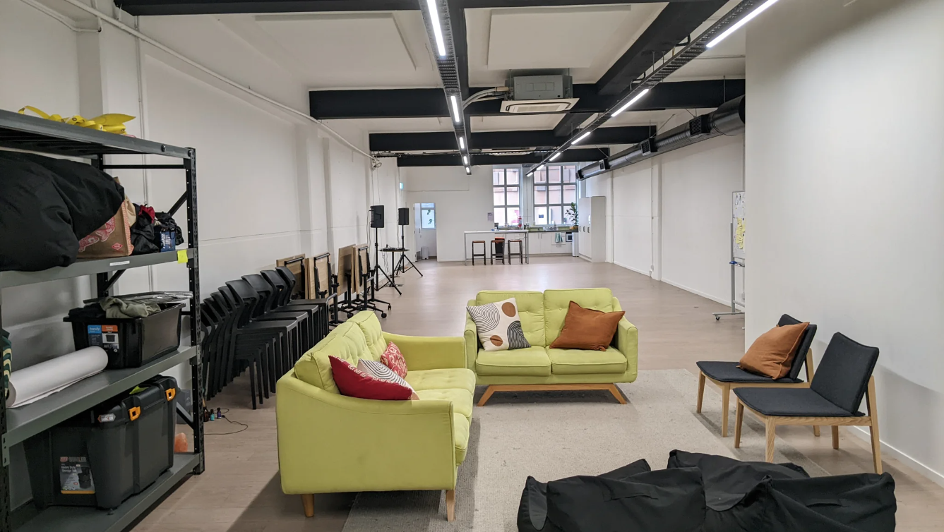 Image Description: A long room with comfy couches in the foreground, a large empty space in the middle and a kitchenette at the end. 