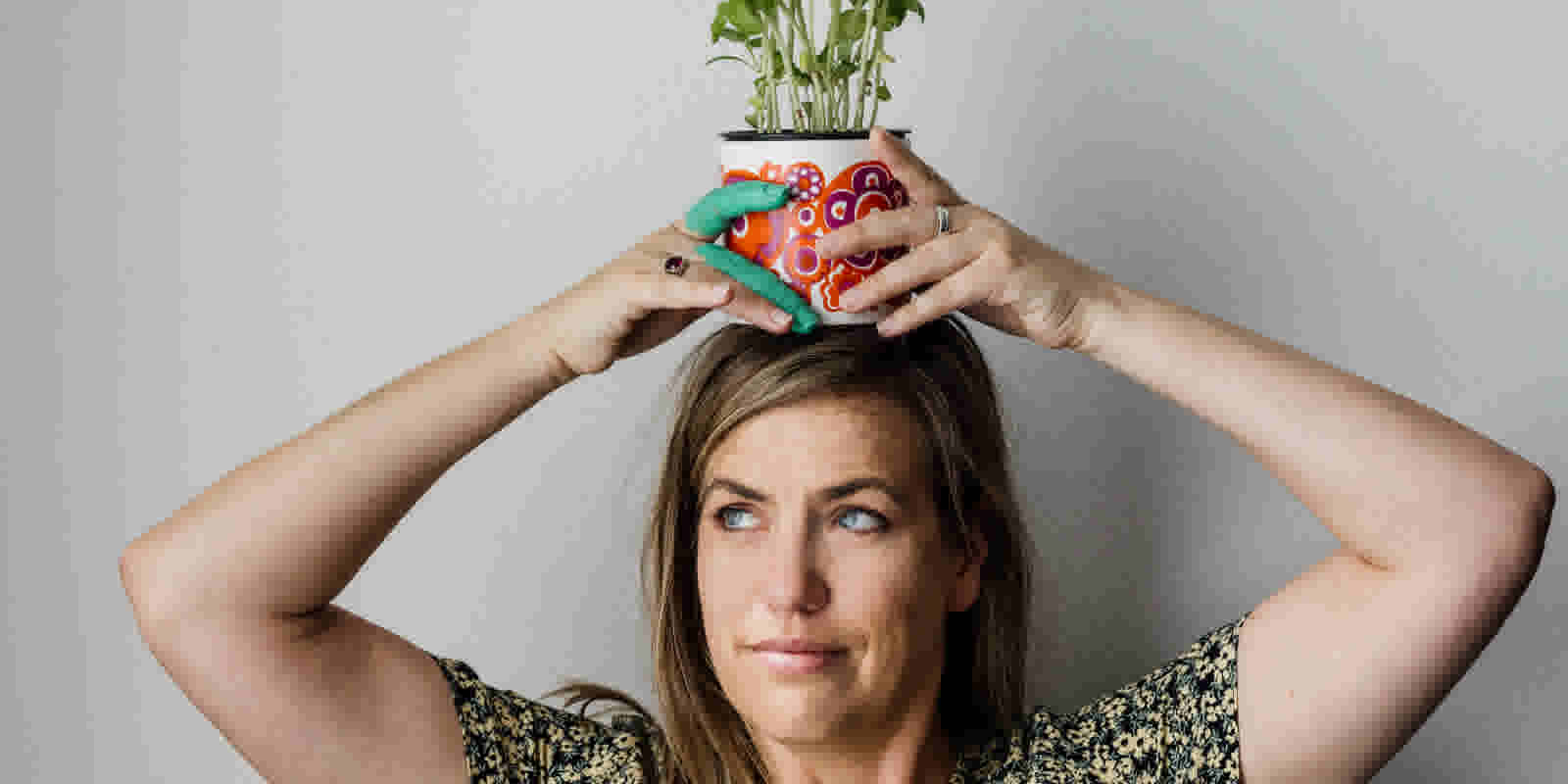 A woman looks to the side while holding a pot plant on her head. Two of her fingers are painted green.