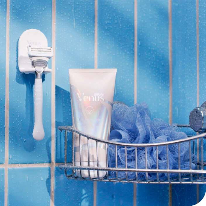 Razor resting on a stand attached to a bathroom wall next to a metal shower basket containing shaving cream and a luffa