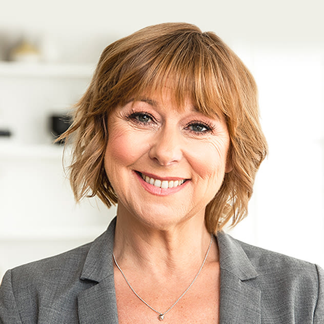 A smiling woman with short, wavy hair wearing a gray blazer is looking at the camera in a bright room.