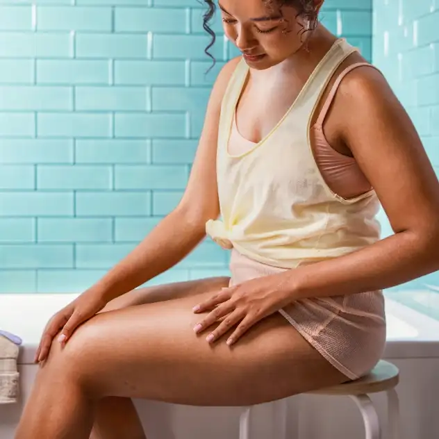 Woman seated on a stool with legs visible beside a bathtub and razor on a folded towel