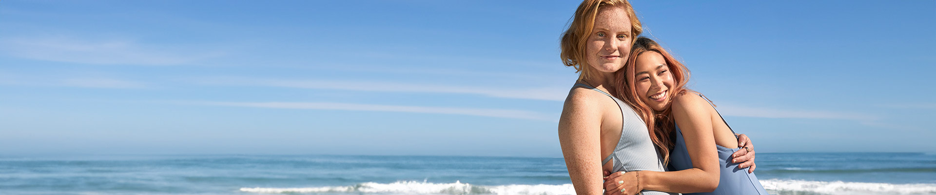 Two young women in swimwear smiling and embracing each other while standing on a beach with ocean waves in the background.