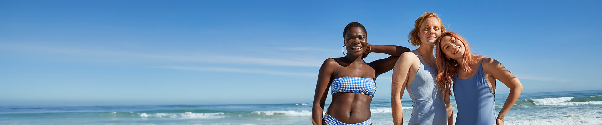 Three women in swimwear smiling together on the beach, with waves in the background and clear blue skies above.