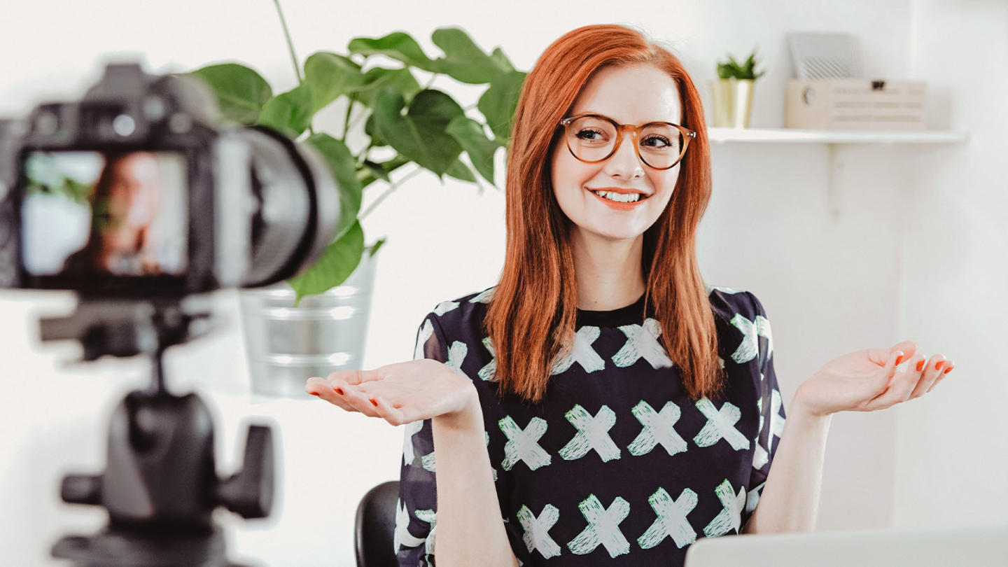 Picture of a redheaded woman in front of a camera with hands raised in a shrugging motion