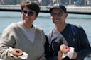 Two people smiling and holding donuts, standing outdoors by a river with a bridge and city buildings in the background. One donut is plain, the other is pink with colorful sprinkles.