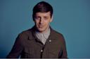 A man with short brown hair wearing a green jacket over a buttoned-up light gray shirt stands in front of a plain blue background, looking directly at the camera with a slight smile.