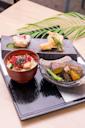 A Japanese meal with a bowl of sashimi over rice, a plate of tempura, and a dish of grilled vegetables, served on a black lacquer tray with a decorative leaf in the background.