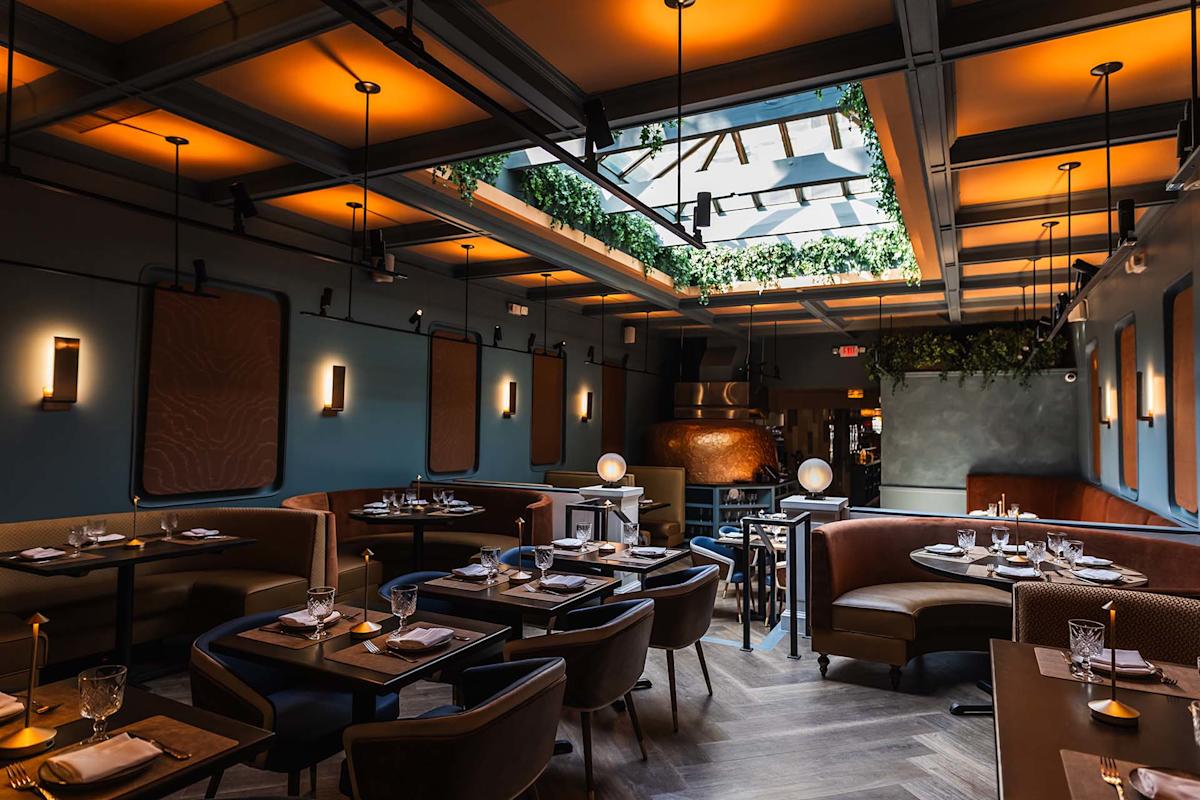 A modern restaurant interior with warm lighting, cozy booth and table seating, a glass ceiling with hanging greenery, and elegant table settings ready for guests.