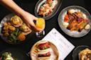 A variety of brunch dishes are displayed on dark plates, including sliders, a meat and fruit platter, and a basket with bao buns. A hand reaches for a glass of orange juice beside a menu titled "Brunch" on the black tablecloth.