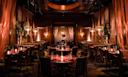 A dimly lit, elegant restaurant with dark wood tables, cushioned chairs, warm pendant lighting, and soft drapes, creating an intimate and upscale atmosphere.
