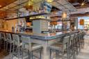 A modern bar with a U-shaped counter surrounded by high chairs. The shelves above are stocked with various bottles. The interior features industrial-style decor with metal accents and warm lighting. Tables and seating are visible in the background.
