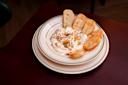 A plate of creamy cheese dip garnished with herbs and red pepper flakes is surrounded by slices of toasted bread on a wooden table.