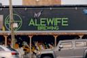 The exterior of Alewife Brewing, with a large green sign on a dark brick wall and murals below. A Jeep and other parked cars are in front, and a "No Parking" sign is attached to a pole.