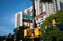 Street signs for E 164 St and Grand Concourse on a pole with traffic lights, trees, and tall apartment buildings against a blue sky in the background.