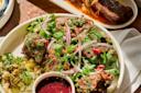 A plate of grilled meat topped with green sauce, fresh cilantro, red onion slices, and pomegranate seeds, served with pickled vegetables and a small dish of red dipping sauce. Another meat dish is blurred in the background.