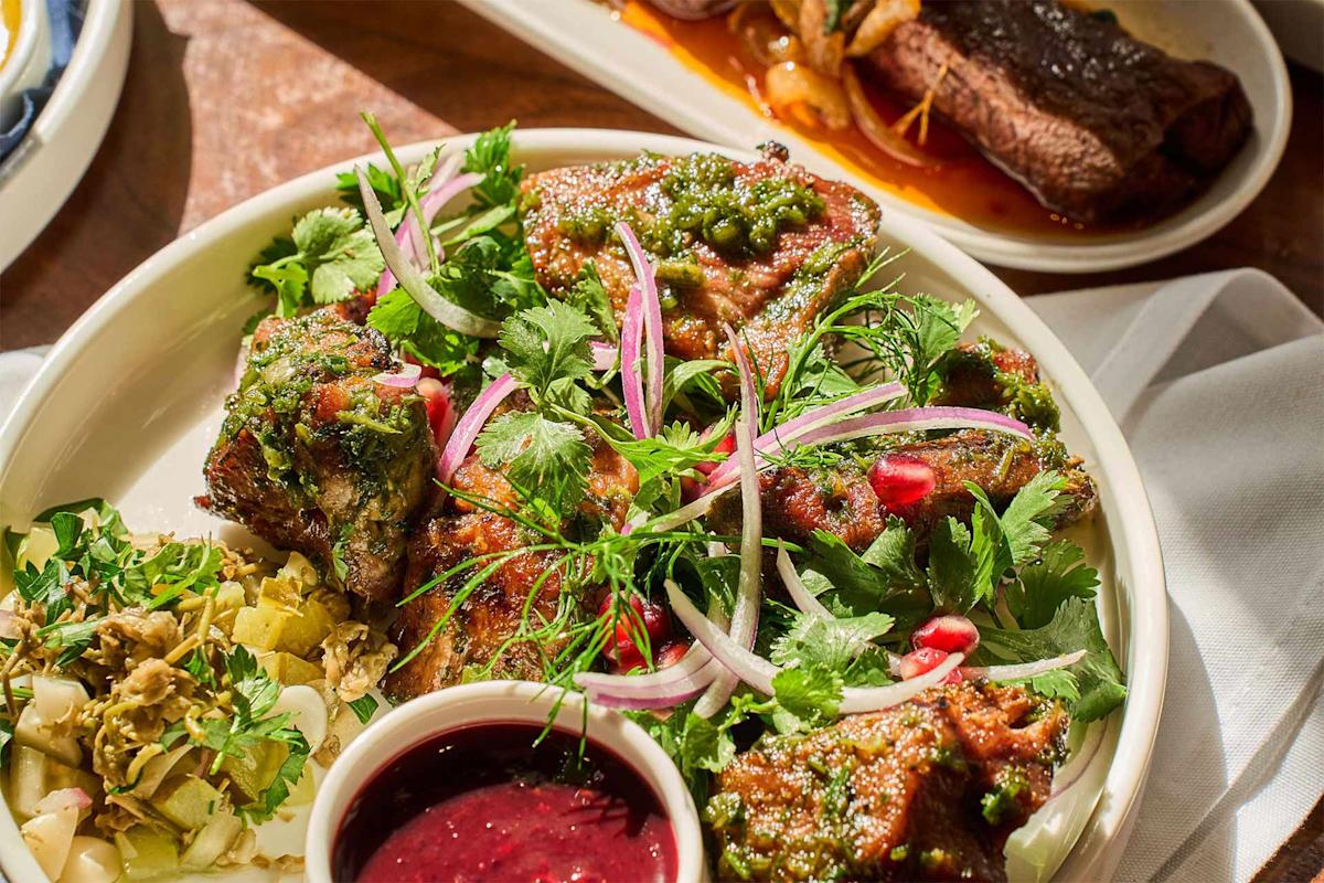 A plate of grilled meat topped with green sauce, fresh cilantro, red onion slices, and pomegranate seeds, served with pickled vegetables and a small dish of red dipping sauce. Another meat dish is blurred in the background.