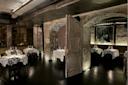 Dimly lit restaurant with exposed brick walls and arched windows. Elegant round tables covered with white tablecloths are set with glassware. A large wooden door is slightly ajar, creating an inviting atmosphere. A black-and-white photo hangs on the wall.