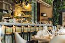 Interior of a modern restaurant featuring a stylish bar with high chairs. Ambient lighting highlights glass bottles and plates on shelves. Tables are set with wine glasses, surrounded by lush greenery on the walls.