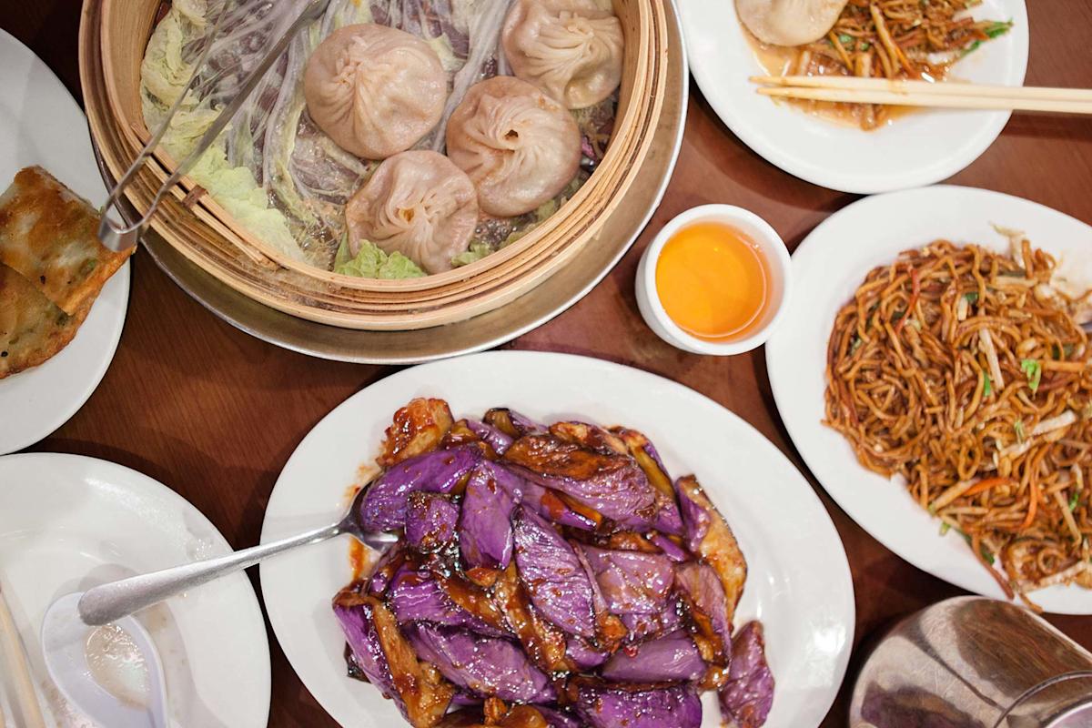 A table set with Chinese dishes, including soup dumplings in a bamboo steamer, stir-fried noodles, braised eggplant, a dipping sauce, and other plates of food on white dishes.
