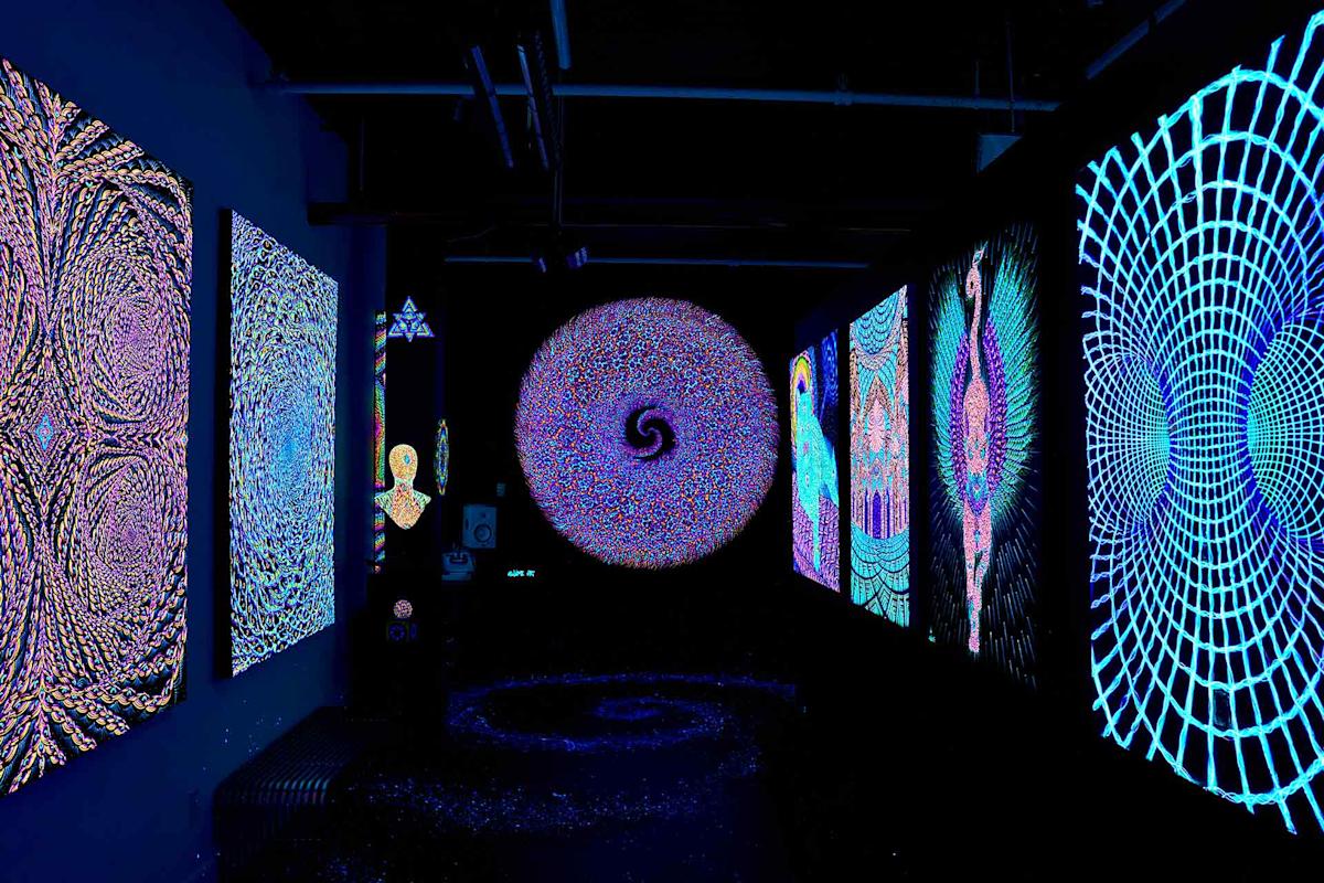 A dark room features glowing, psychedelic art on the walls, including intricate patterns, mandalas, a central circular art piece with a spiral design, and vibrant blue, purple, and neon colors throughout.