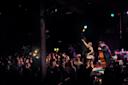 A woman sings and gestures on stage with a band, including an upright bass player and accordionist, while a large audience watches in a dim, intimate music venue.