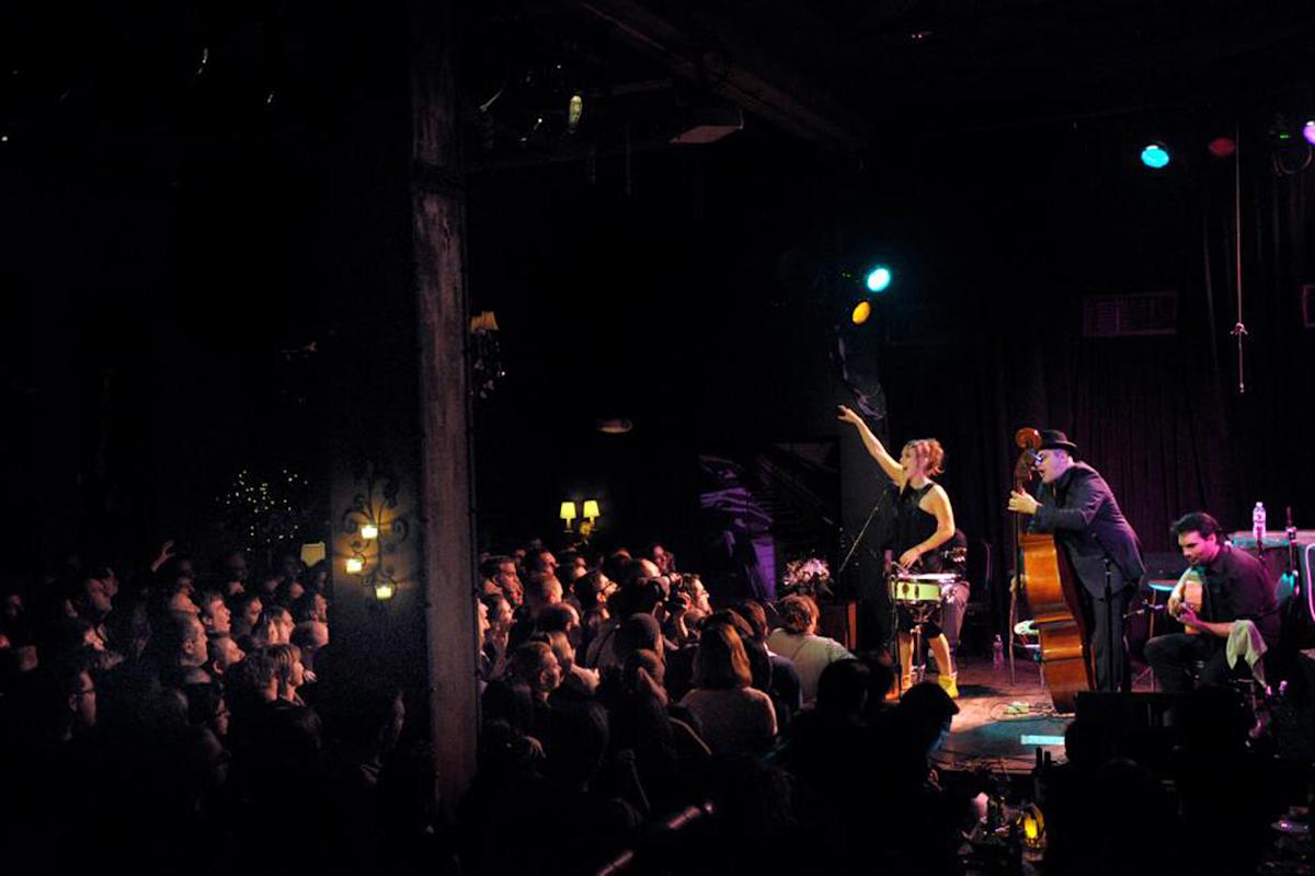 A woman sings and gestures on stage with a band, including an upright bass player and accordionist, while a large audience watches in a dim, intimate music venue.