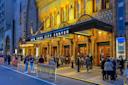 People gather outside the illuminated New York City Center theater at dusk, standing in line near ticket barriers on the street, under the ornate marquee and decorative facade.