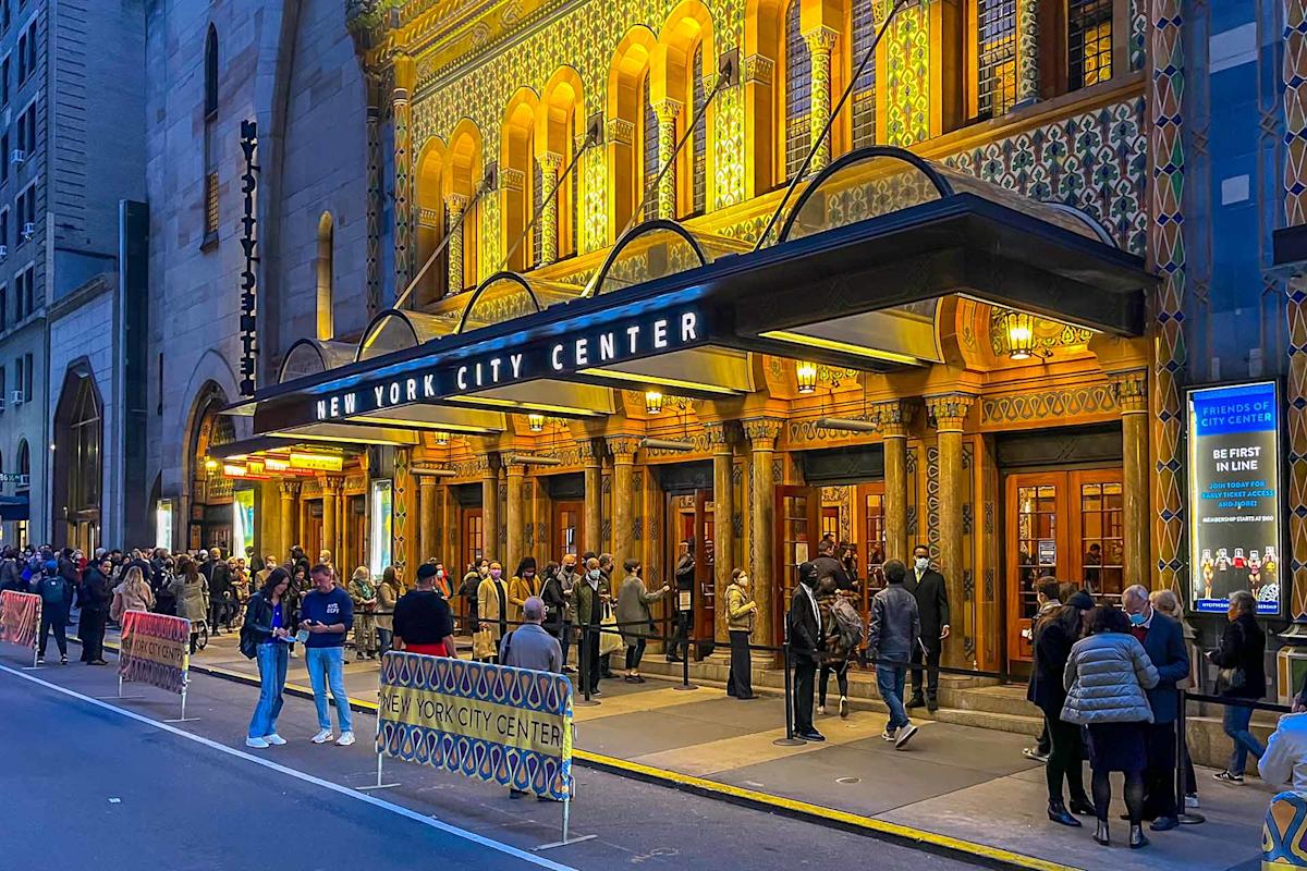 People gather outside the illuminated New York City Center theater at dusk, standing in line near ticket barriers on the street, under the ornate marquee and decorative facade.