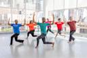 A group of six adults in colorful shirts and black pants practice a dance routine in a bright studio with large windows overlooking city buildings. They pose with arms raised and bent knees, focused and synchronized.